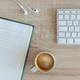 Minimalistisch bureau met wit toetsenbord, notitieblok, kopje koffie en witte oordopjes op houten tafelblad