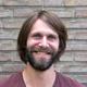 Een glimlachende man met halflang donker haar en een baard, gefotografeerd tegen een bakstenen muur achtergrond