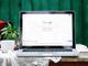 Laptop met Google-startpagina op scherm, naast rode koffiepot en witte kop op rustieke houten tafel met kanten kleed