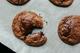 Vers gebakken chocoladekoekjes op bakpapier, één koekje met een hapje eruit waaruit gesmolten chocolade zichtbaar is.