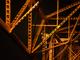 Detail van een stalen brugconstructie 's nachts verlicht in warme oranje tinten, met geometrische patronen van balken en spanten.