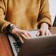 Persoon in warme gele trui werkt op laptop aan houten bureau, close-up van handen met horloge.