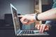 Hand met fitnesstracker wijst naar laptopscherm tijdens gebruik, close-up van interactie met touchpad en toetsenbord