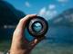 Hand houdt cameralens vast met bergmeer in focus, waarbij het landschap door de lens wordt weerspiegeld tegen blauwe lucht
