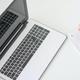 Hand in grijze trui schrijft in notitieblok naast opengeslagen laptop op wit bureau, bovenaanzicht