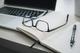 Leesbril op open notitieboek naast laptop en pen op wit bureau, moderne werkplek opstelling