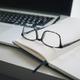Leesbril op open notitieboek naast laptop en pen op wit bureau, moderne werkplek opstelling
