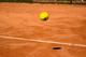 Helgele tennisbal zweeft boven een rode gravel tennisbaan met schaduw eronder, witte lijn zichtbaar