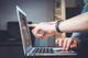 Hand met fitness tracker wijst naar laptopscherm tijdens online werksessie op een donker bureau.