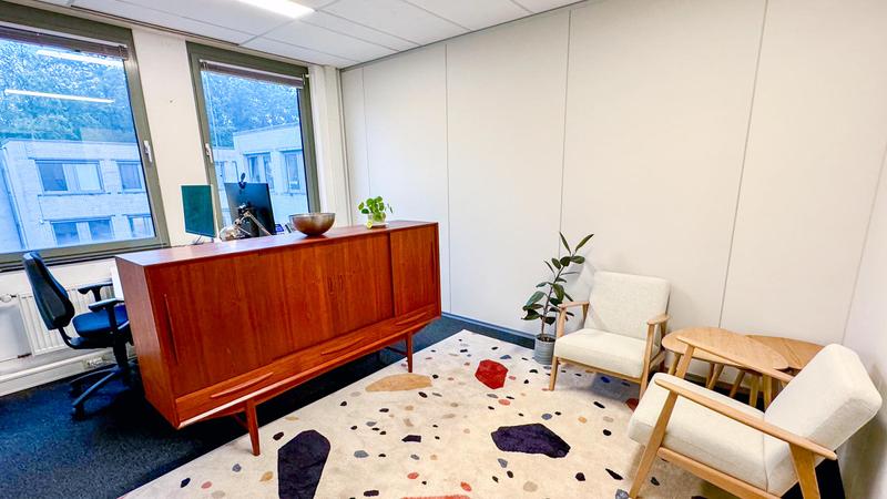 Modern kantoorruimte met vintage dressoir, witte fauteuils en terrazzo-print tapijt naast grote ramen met daglicht.
