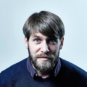 Professioneel portret van een persoon met baard en blauwe ogen in een donkerblauwe trui tegen een lichte achtergrond.